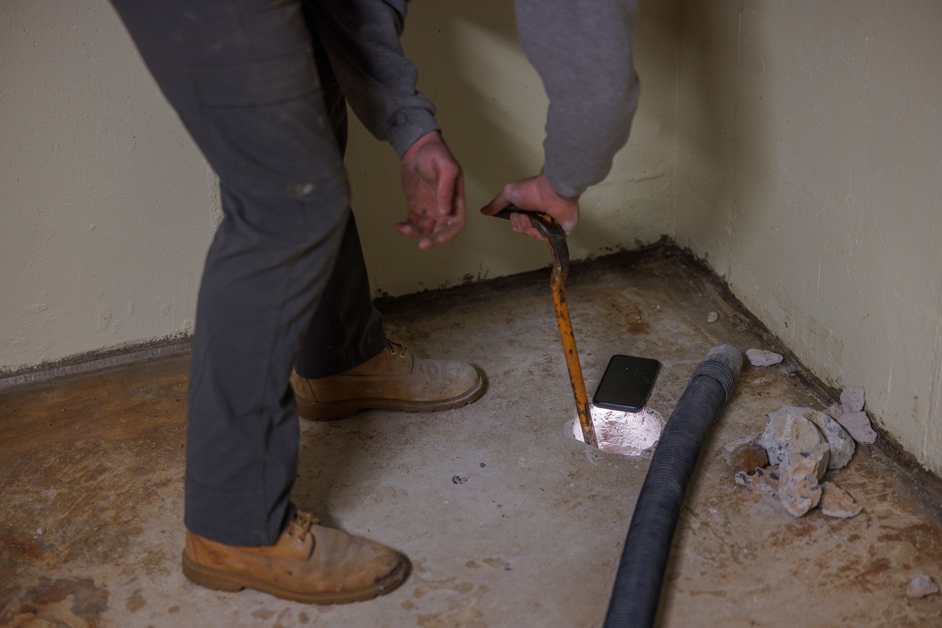 A worker is making a hole in the basement floor to install a radon mitigation system, using a crowbar.